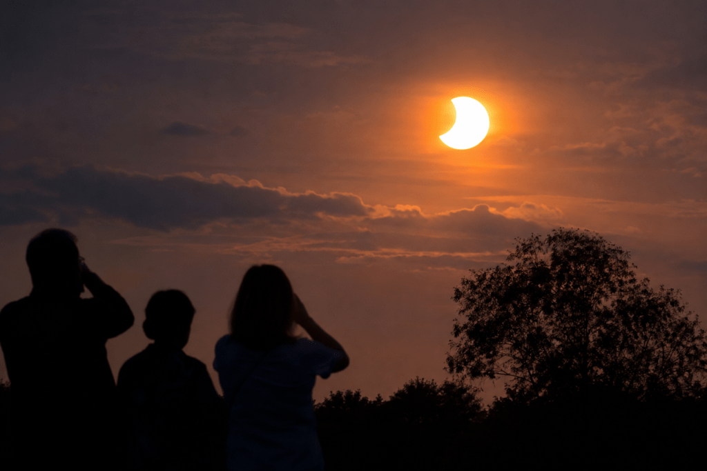 Silhouettes of three people watching a solar eclipse against a colorful sunset sky with clouds.