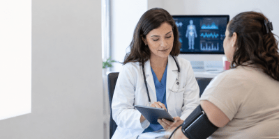 Medica consulta una paziente durante una valutazione clinica con misurazione della pressione e dati sanitari visualizzati sullo sfondo.