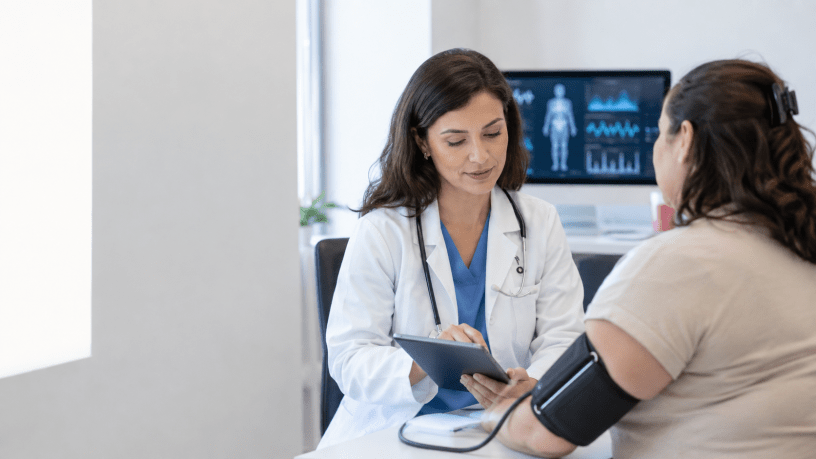 Medica consulta una paziente durante una valutazione clinica con misurazione della pressione e dati sanitari visualizzati sullo sfondo.