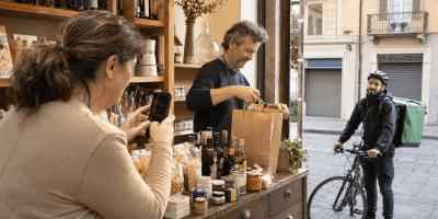 Due commercianti preparano una consegna in un piccolo negozio mentre una donna fotografa i prodotti con lo smartphone e un ciclista attende all’ingresso.