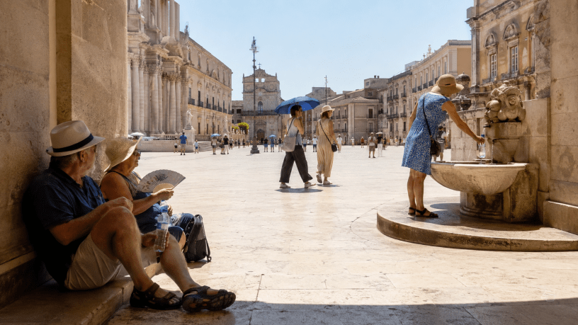 Persone cercano riparo dal caldo e si riforniscono d’acqua in una piazza italiana assolata durante un’ondata di calore estremo.