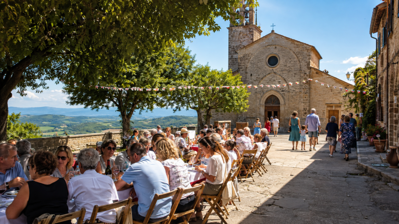 Famiglie e amici riuniti per il pranzo di Ferragosto nella piazza di un borgo italiano, davanti a un’antica chiesa dedicata alla tradizione dell’Assunta.