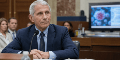 Rappresentazione realistica di Anthony Fauci seduto davanti a un microfono in un’aula istituzionale americana, con sullo sfondo un’immagine del coronavirus.