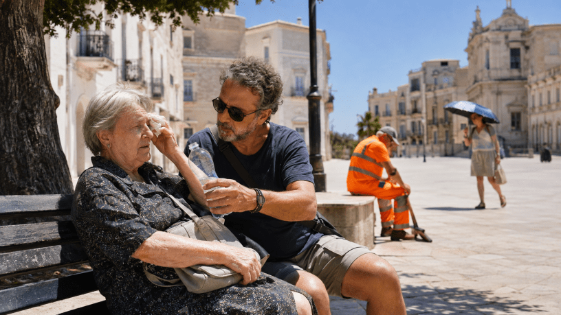 Un uomo offre una bottiglia d’acqua a una donna anziana provata dal caldo, seduta all’ombra in una piazza italiana durante un’ondata di calore.