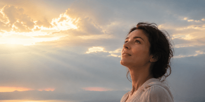 : Donna con lo sguardo rivolto verso il cielo, illuminata dai raggi dorati del sole tra le nuvole, immagine ispirata alla poesia “Serafico” di Francesca Giordano.