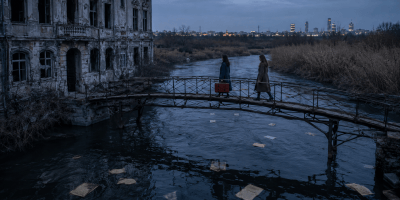 Due donne attraversano un vecchio ponte sopra un fiume, mentre documenti abbandonati galleggiano accanto a un edificio in rovina.