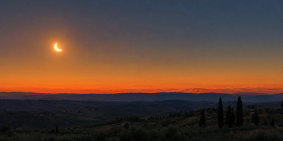 Eclissi parziale di Sole al tramonto sopra le colline italiane, con il disco solare ridotto a una falce luminosa.