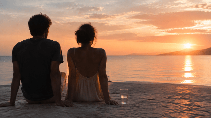 Due persone sedute di fronte al mare al tramonto, con un bicchiere d’acqua accanto.