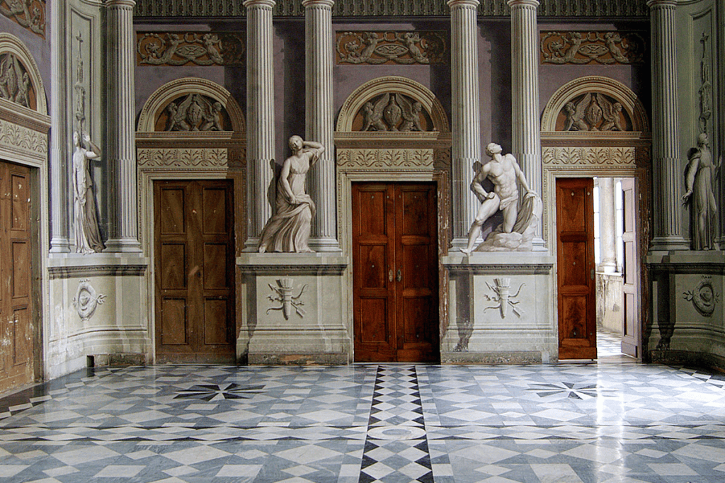 Interior view of a classical hall featuring decorative columns, sculptures of figures, and wooden doors. The floor has an intricate geometric pattern.