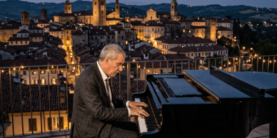 Man in suit playing grand piano on stone terrace with lit historic town in background at dusk
