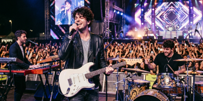 Rock band The Kolors performing on stage with guitar, drums, and keyboard in front of a large audience