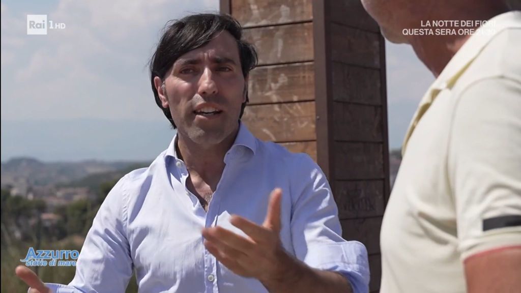A man with dark hair wearing a light blue shirt gestures while speaking, with a scenic view in the background.