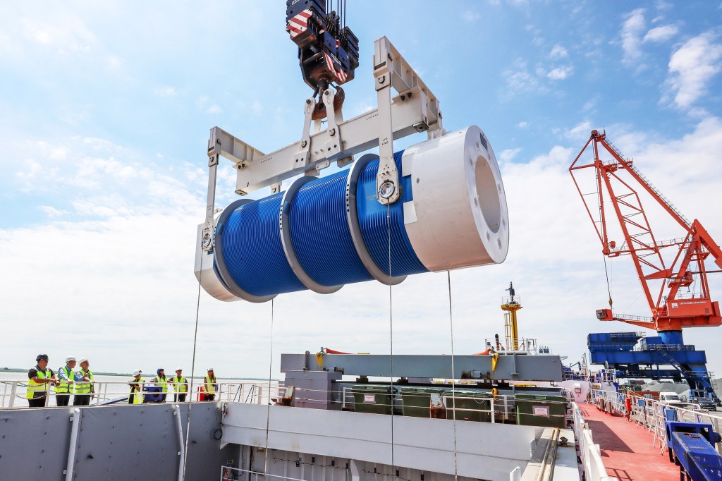 A large blue and white spool being lifted by a crane on a cargo ship, with workers observing from the deck.