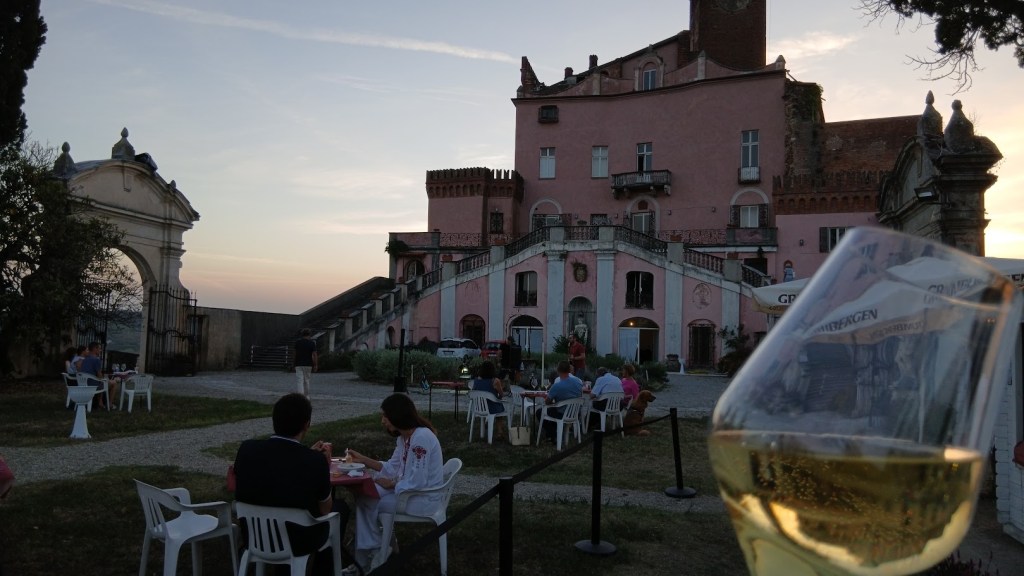 An outdoor dining scene at a pink castle with a glass of white wine in the foreground. People are seated at tables enjoying their meals as the sun sets in the background.