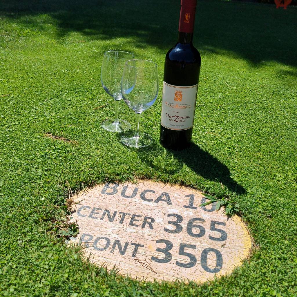 A bottle of red wine and two wine glasses on green grass, beside a marker displaying 'BUCA 10 CENTER 365 PONT 350'.