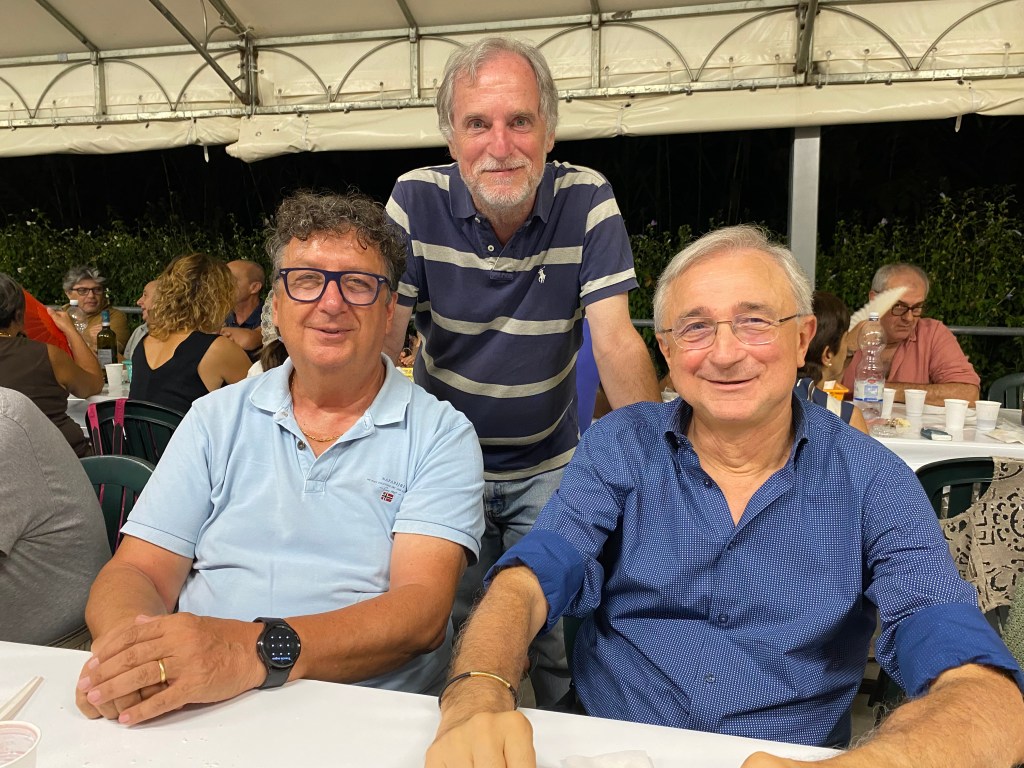 Three men sitting at a table in a festive outdoor setting, with one standing behind them. They are smiling and enjoying a social gathering.