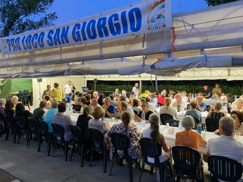 A large gathering of people seated at long tables under a canopy, enjoying a meal during an evening event, with a banner reading 'Pro Loco San Giorgio' in the background.