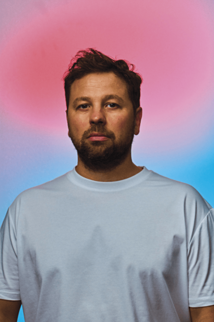 A man with a beard and short hair, wearing a plain white t-shirt, stands in front of a softly illuminated background featuring hues of pink and blue.