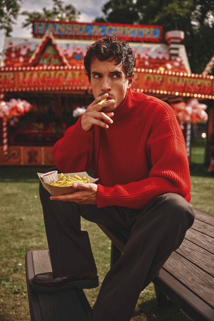 A young man in a red sweater sitting on a wooden bench, holding a plate of fries in one hand and a cigarette in the other, with a colorful carnival backdrop featuring the words 'Hansel & Gretel'.