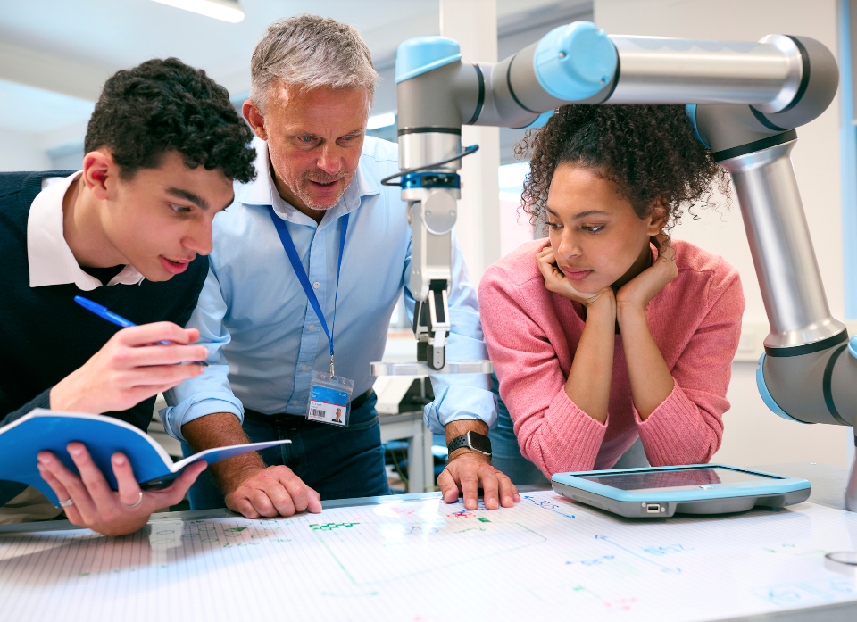 Three individuals collaborating in a technology lab, examining a robotic arm and discussing notes while engaging with a tablet.