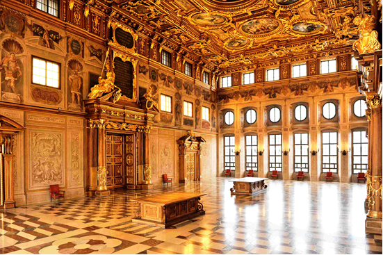 An opulent grand hall featuring ornate golden ceilings, intricate wall designs, large windows, and a polished checkered floor.