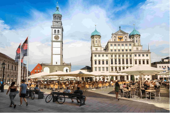 A lively square with outdoor dining, surrounded by historical buildings including a tall clock tower and a large ornate structure with domed roofs, under a blue sky with scattered clouds.