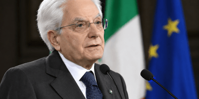 Sergio Mattarella interviene davanti ai microfoni con le bandiere dell’Italia e dell’Unione Europea sullo sfondo.