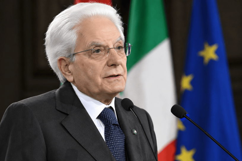 Sergio Mattarella interviene davanti ai microfoni con le bandiere dell’Italia e dell’Unione Europea sullo sfondo.