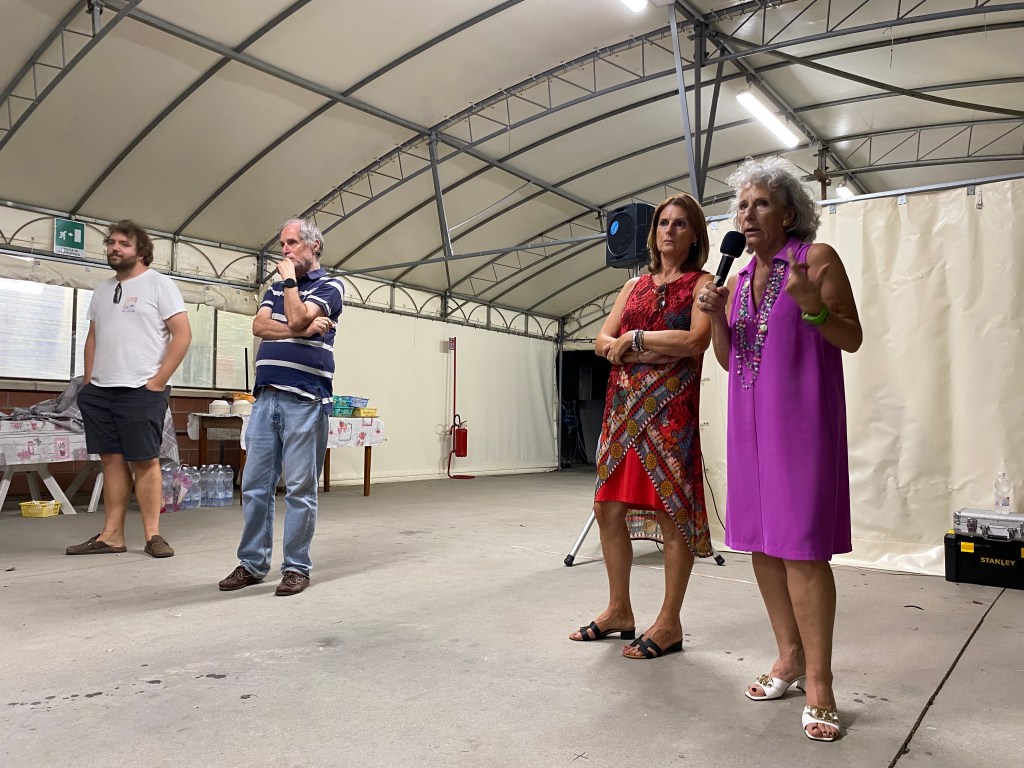 A group of four people inside a large tent-like structure, with two women speaking into a microphone while the others listen. One woman is wearing a pink dress and the other a red patterned dress. The men are standing nearby, appearing engaged in the discussion.