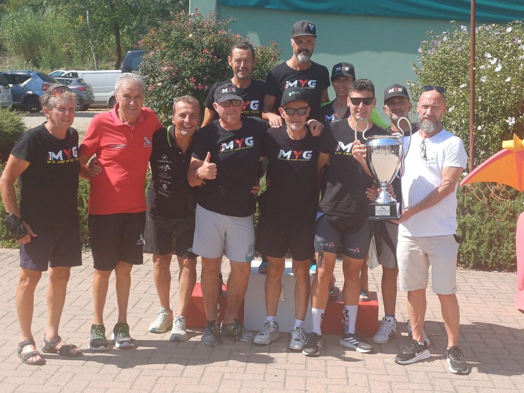Group of ten people celebrating with a trophy outdoors, wearing matching shirts with the MYG logo, in a sunny setting.