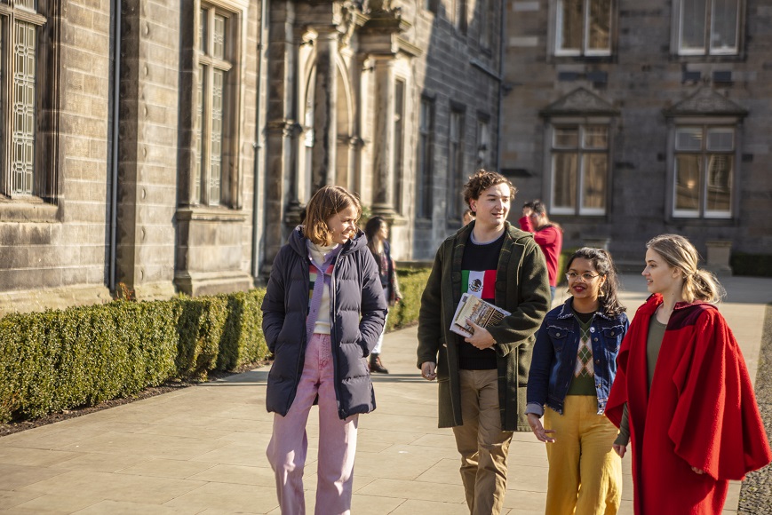 A group of four young adults walking together on a sunny day in front of a historic building. They are engaged in conversation and wearing casual clothing.