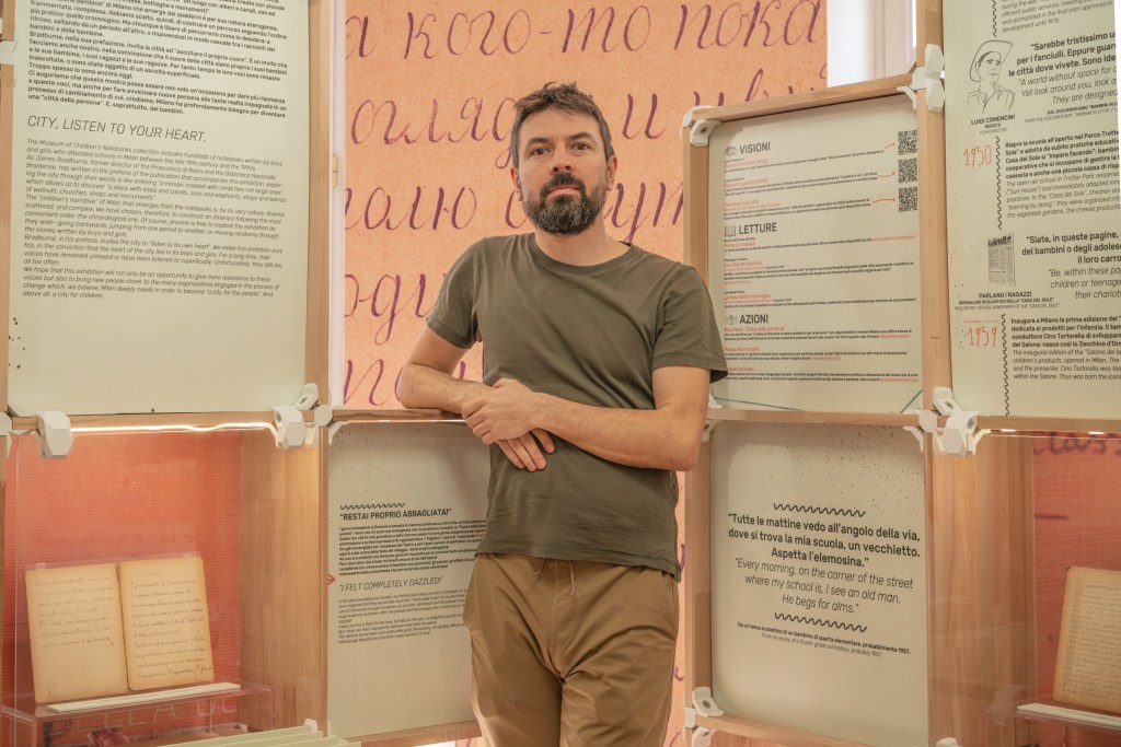 A man standing in front of display panels featuring text and illustrations related to urban themes, with a friendly expression and casual attire.
