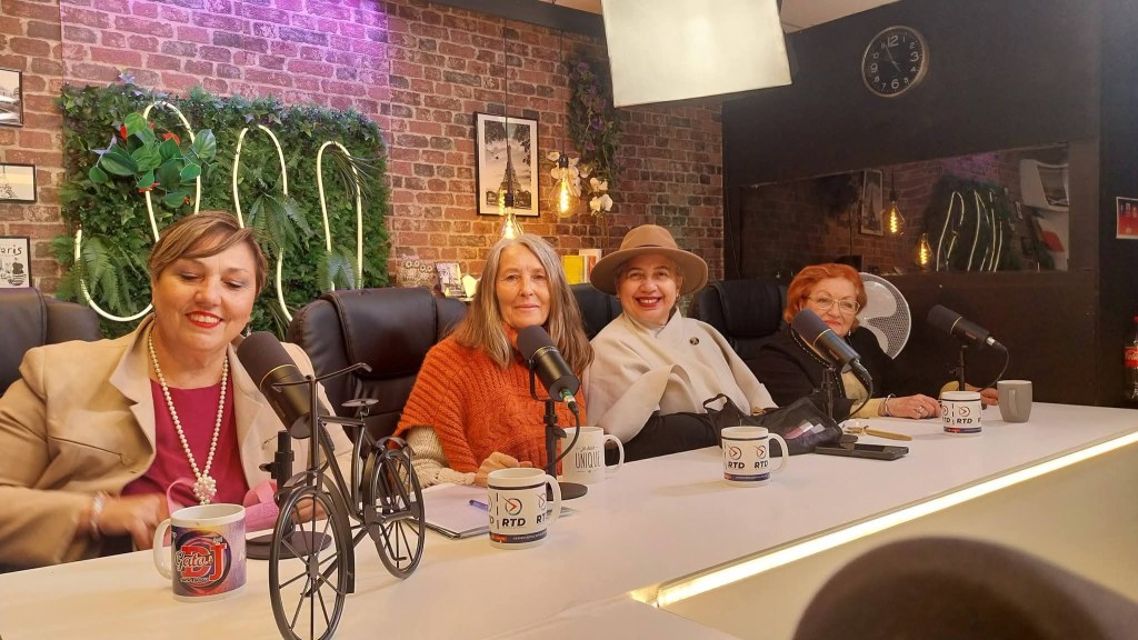 A group of four women seated at a podcast studio table, with microphones and coffee mugs in front of them. The backdrop features brick walls, plants, and decorative elements.
