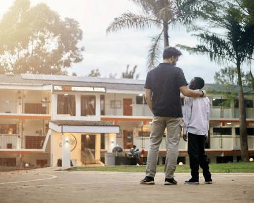 An adult and a child standing together in front of a school building, with trees in the background.