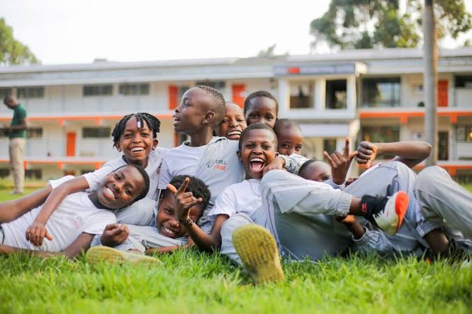 A group of happy children lying on grass, smiling and posing for the camera. They are wearing matching gray t-shirts and appear to be enjoying a fun moment outdoors.