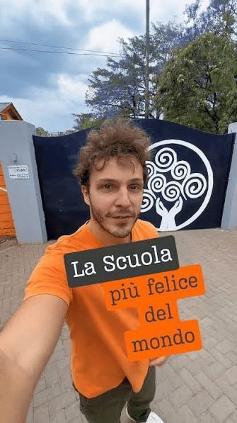 A young man with curly hair wearing an orange shirt takes a selfie in front of a large painted wall featuring a tree logo and the text 'La Scuola più felice del mondo' in Italian, which translates to 'The happiest school in the world'.
