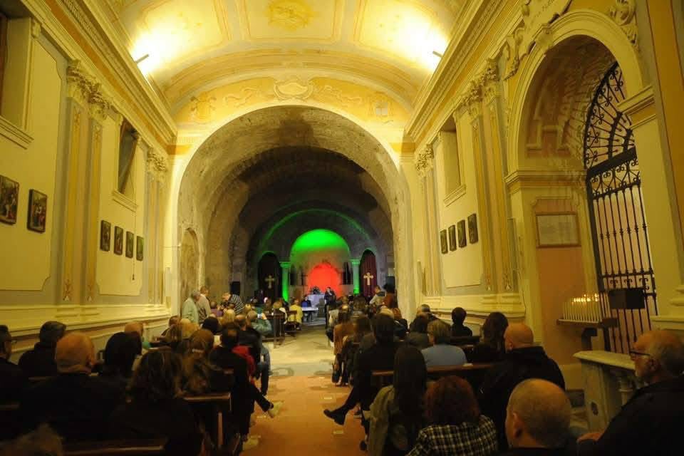 An audience seated in a dimly lit church or hall, facing a stage with performers under colorful lighting. The decor features arched ceilings and ornate walls.
