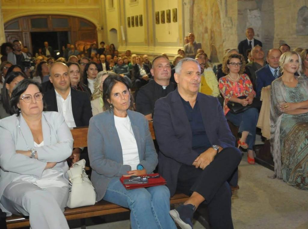 A group of people seated in an auditorium, engaged in a presentation or event. Some individuals appear focused, while others are chatting or looking around the room.
