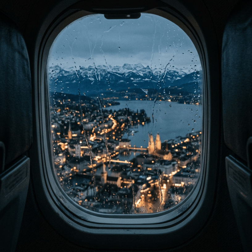 Rain-speckled airplane window overlooking a lakeside city and snow-capped mountains